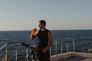 Obraz premium Handsome young man drinking water and leaning at his bicycle with sea on the background