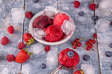 Balls of homemade strawberry, red currant, raspberry, blueberry ice cream, sorbet in bowl, scoops, berries and ice cubes over on blue wooden background.  summer dessert, sweet snack.