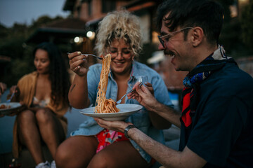 Loving couple sitting outdoors in house yard during an evening, and sharing a plate of pasta spaghetti.