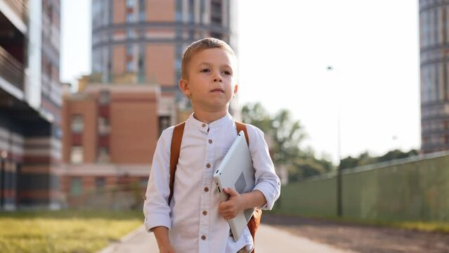 Caucasian boy goes to school in full dress, in white shirt with backpack. Child left house and walks yard. Preparation for school. Preschooler became first grader. Knowledge day. Getting an education.