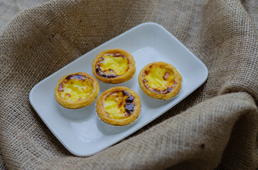 Homemade small egg tarts on a white plate, dessert Portuguese. on brown fabric background.