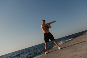 Fototapeta na wymiar Full length of muscular man doing stretching exercises outdoors with the sea on background