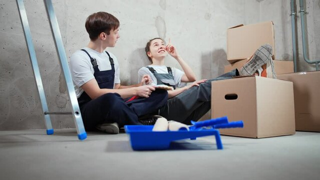 Young married couple sit in room communicate, discuss, laugh and smile, chooses color for design and interior. Home renovation and comfort. In foreground paint roller, repair tools and concrete floor.