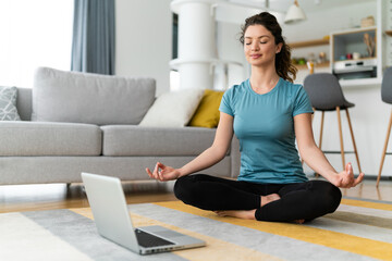 Young woman with ponytail doing yoga and meditating while listening to relaxed music on laptop at living room.