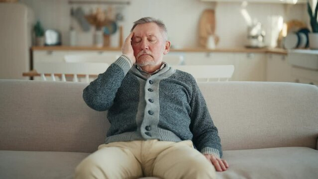 Senior man suffering from terrible headache touching head sitting on couch at home, making massage with closed eyes,doing turning head on sides exercise. Migraine, high intracranial pressure concept.