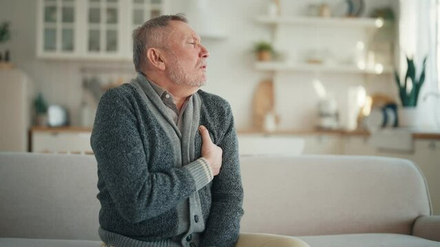 Senior man suffers from heartache sitting on couch at home. Unwell old male with chest pain myocardial infarction has chronic cardiology disease. Cardiac arrhythmia, heart attack, illness concept.
