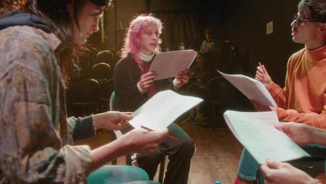Group of young actors sitting in circle on stage, reading play scripts printed on papers during rehearsal with male theater director