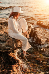 woman sea sunset. woman in a white pantsuit and hat is sitiing on the beach enjoying the sea. Happy summer holidays
