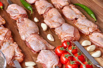Metal skewers with raw uncooked chicken meat for frying on the wooden board with peppers near it, close-up perspective view. Shallow depth of field. Chicken and garlic in focus