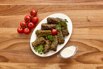 White plate with dolma (meat in grape leaves) on the wooden table with red cherry tomatoes, garlic, greens and sour cream near it. Top view, flat lay