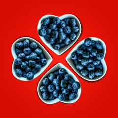 Creative summer mosaic of blueberries in white heart shaped bowl on vibrant red background. Original blueberry decoration. Minimal summer concept. Welcome summer. Copy space. Clover shape pattern.