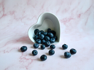 Creative summer layout of blueberries and white heart shaped bowl on pastel pink background. Original blueberry decoration. Minimal summer concept. Welcome summer. Copy space.