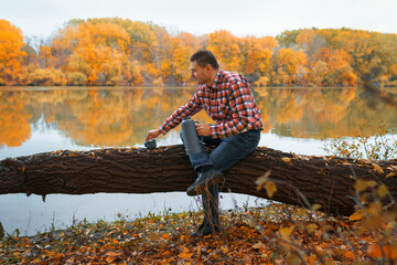 a man sits on a log on the river bank and drinks tea or coffee from a thermos, enjoys the landscape, beautiful nature in the autumn season