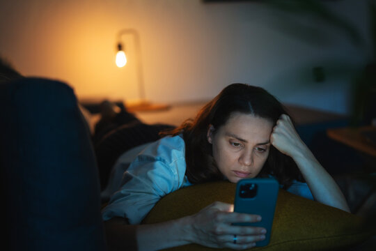 Young Serious Sad Woman Lying On Sofa And Looking At Smartphone Screen At Night Time At Home