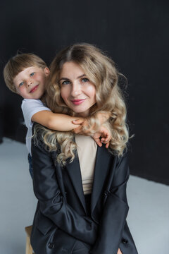 Mom and son at a photo shoot in the studio. Mom is wearing a black pantsuit. The boy is wearing a white T-shirt. The son hugs his mother by the neck. The background in the photo is black and white.