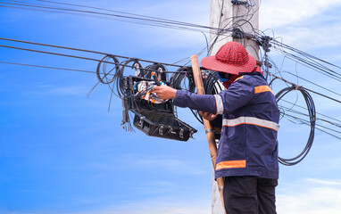 Technician on wooden ladder checking fiber optic cable lines in splitter box while repairing internet connection system on electricity pole against blue sky background