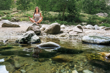 The beautiful girl practices Yoga on the bank of the mountain river. The young woman is sitting on a stone by the water's edge.