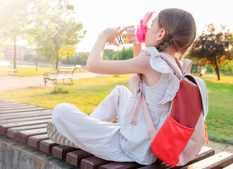 Little girl with a backpack sits on a bench and drinks water from a bottle in the park. Back to...
