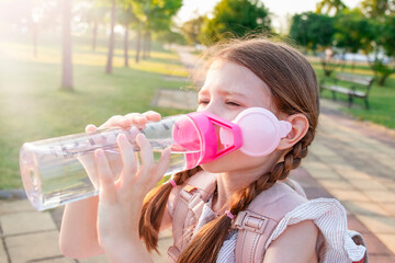 Little girl with a backpack drinks water from a bottle in the park