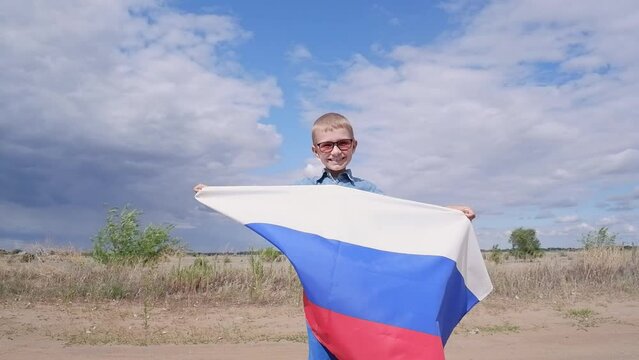 Portrait of a cute, smiling happy boy in a field with a Russian flag on his shoulders. National holiday independence day. Pride and patriotism.