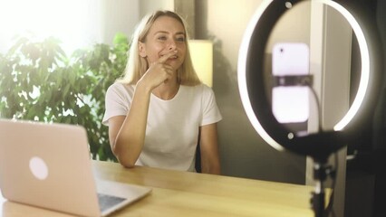 Cheerful young blond woman influencer recording video blog mobile vlog podcast live stream share experience with audience on smartphone using tripod at light home indoors