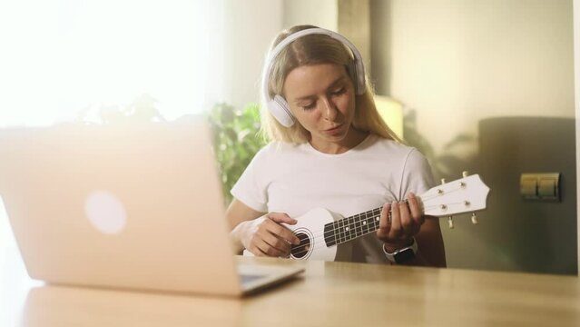 Portrait of pretty young blond woman watch video tutorial on ukulele guitar playing or having personal lesson with teacher distance remote on laptop computer at home Online musical education concept 