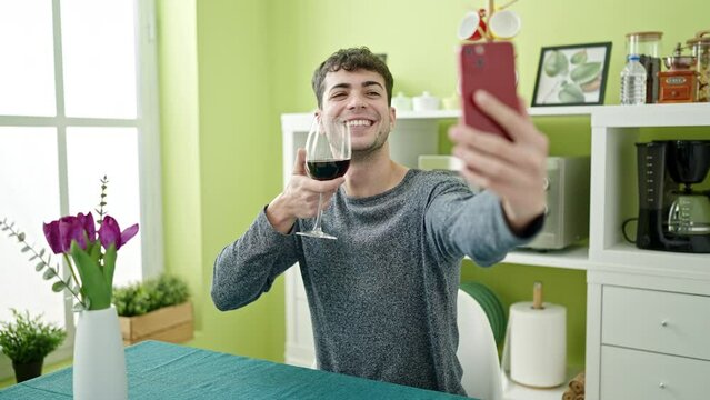Young hispanic man drinking glass of wine taking a selfie with smartphone at dinning room
