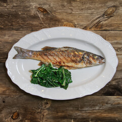 Grilled fish Seabass with spinach garnish served on a white plate on a wooden rustic table