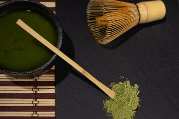 bowl of black matcha tea with gray stains whisk and bamboo spoon on table with cane tablecloth with green tea old cement background with stains close-up top view photo with space for text 