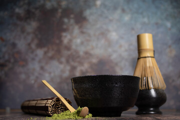 tea matcha black bowl with gray stains whisk and bamboo spoon on wooden table with tea green powder on top old cement type out-of-focus background and a rolled cane tablecloth horizontal close-up phot