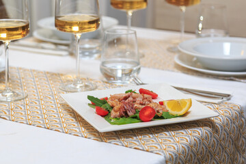 square plate with mini calamari and garnish of green leaves and cherry tomatoes served with a knife and fork with a glass of water and wine