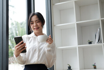 Happy and relaxed Asian businesswoman having a coffee break during the meeting, holding smartphone in her hand.