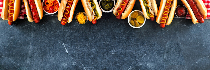 Group of hot dogs with an assortment of toppings and condiments. Overhead view top border on a dark stone background.