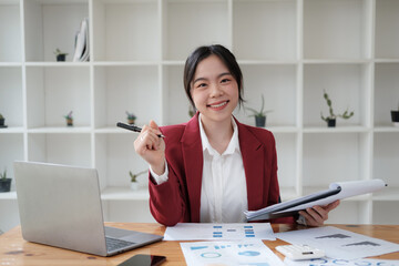 Confident young Asian office worker in red suit working with laptop computer in her office room.