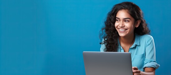 Smiling Indian teen girl using laptop for online study on blue background Happy student in educational webinar