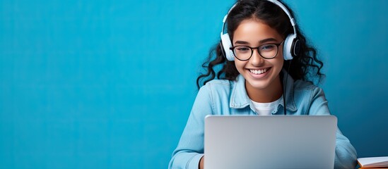 Smiling Indian teen girl using laptop for online study on blue background Happy student in educational webinar - Powered by Adobe