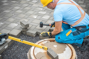Laying interlocking paving. A worker is placing interlocking paving stones around the sewer manhole cover. - obrazy, fototapety, plakaty