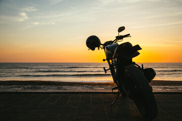 Classic Motorrad am Strand bei Sonnenuntergang im Gegenlicht mit Horizont, Sonne, Meer und Wellen im Hintergrund und Helm am Motorrad, Picknick, Pause, Auszeit, Lebensziel, Bucket List, 