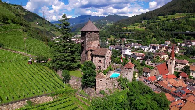 Aerial orbit of medieval castle in Chiusa South Tyrol. Drone flying over garden pool, iconic church and red roofs with vineyards in Italy. Klausen village and Alpine landscape, 4k stock footage.