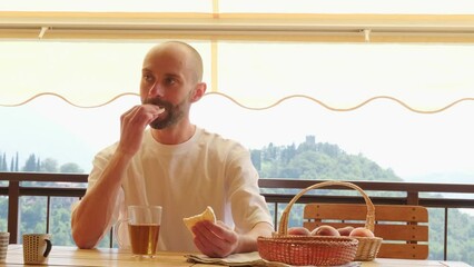 young man 30-35 years old drinks tea from glass cup and eats rice cookies, tea drinking on balcony, loggia flooded with warm light, relaxation in Italy