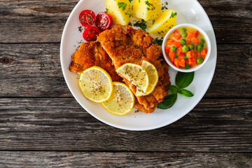 Crispy breaded seared chicken cutlet with boiled potatoes and vegetables on wooden table
