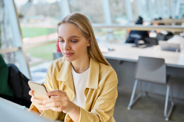 Teen girl gen z student using mobile phone looking at smartphone sitting at desk in university college campus classroom. Young blonde woman holding cellphone technoogy in university.