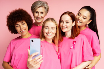 Smiling multiracial women wearing  t shirts with breast cancer pink ribbon holding mobile phone,...