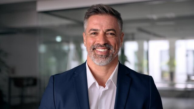 Headshot close up portrait of indian or latin confident mature good looking middle age leader, ceo male businessman in suit on office background. Handsome hispanic senior business man smile at camera.
