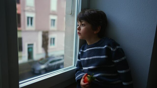 Bored sad emotion of little boy sitting by window staring at view in melancholy, child wanting to go out stuck at home