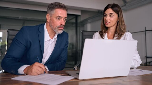 Mature 50s age Latin business man mentoring mid age European business woman discussing project on laptop in office. Two colleagues, group of partners, professional business people working together.
