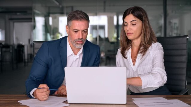 Team of diverse partners sitting at table mature Latin business man and European business woman discussing project on laptop in office. Two colleagues of professional business people working together.