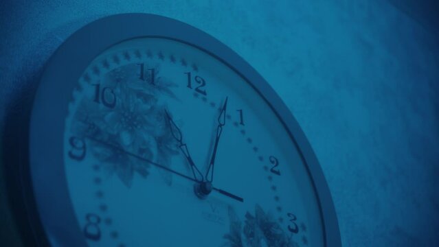 Close-up of wall clock that shows 11 pm in a room under blue light with a smooth second hand. Flowers are depicted on the dial. Evening time