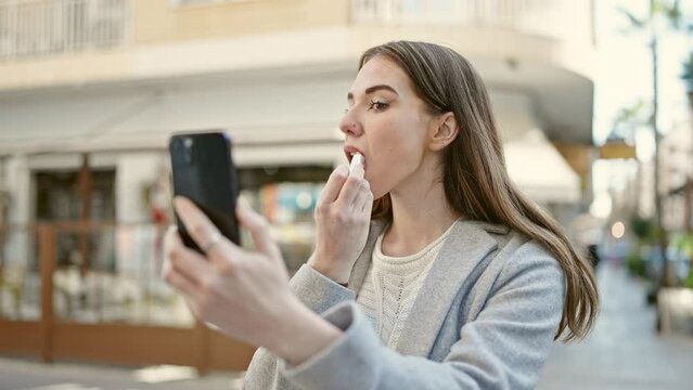 Young hispanic woman using smartphone as a mirror make up lips at coffee shop terrace