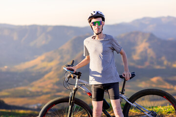Fototapeta premium ortrait of a cyclist resting atop his mountain bike, wearing sunglasses and a helmet, with a mountain landscape in the background